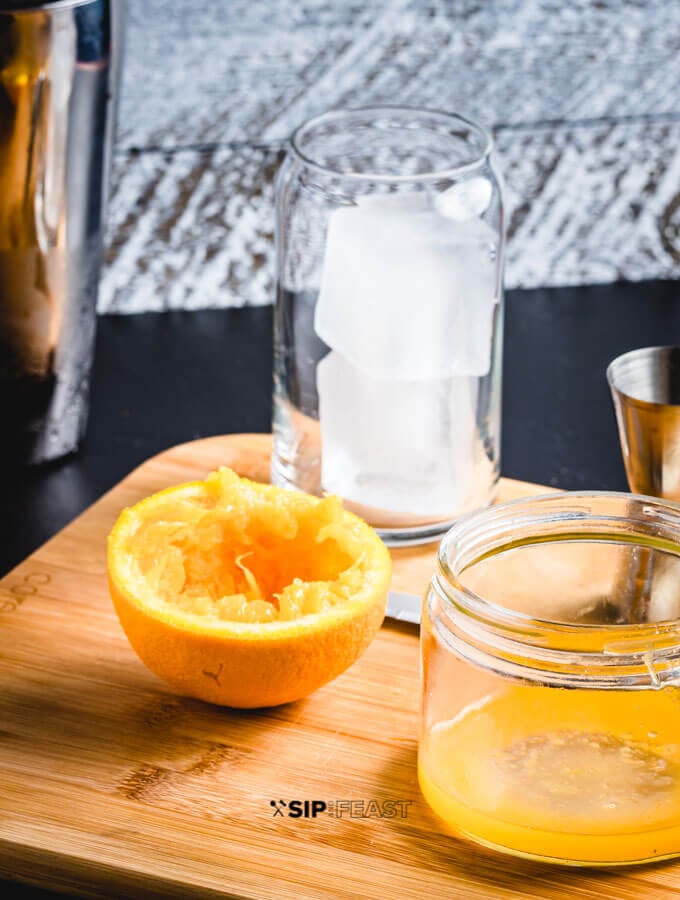 A glass of ice, a cocktail jigger, fresh squeezed orange juice in a citrus juicer and half an orange.