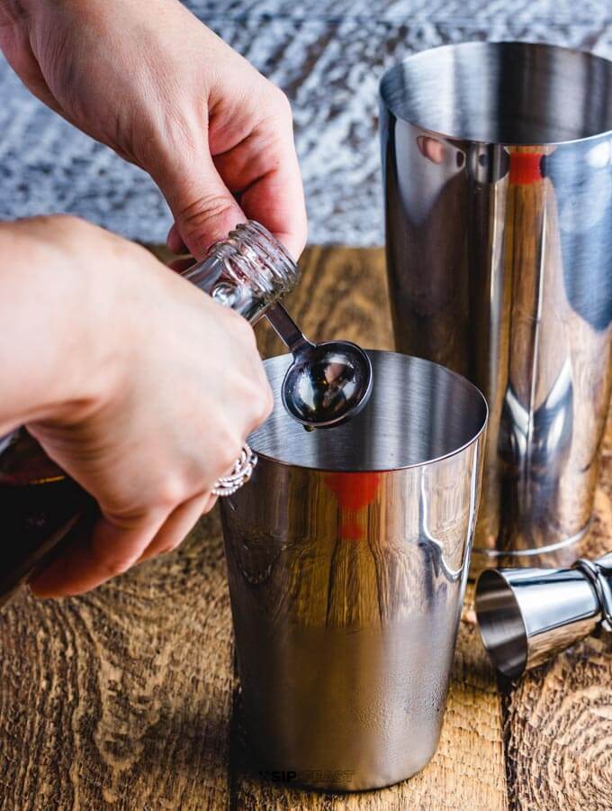 Hands measuring a teaspoon of maple syrup and pouring into a cocktail shaker.