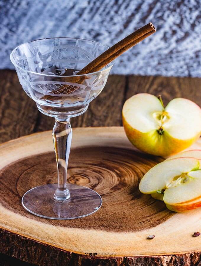 Cocktail glass with cinnamon stick, and sliced apples on a cutting board.