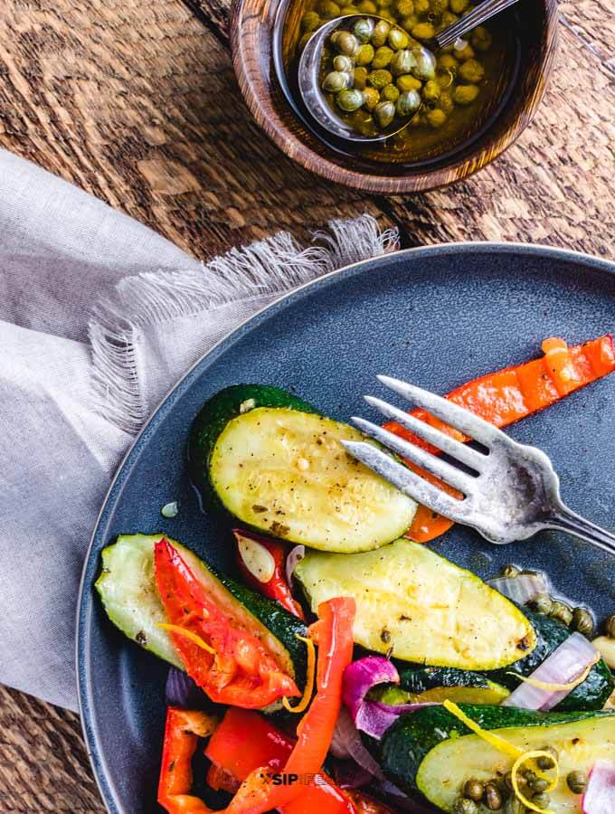 Grey plate with roasted zucchini and peppers along with small bowl of lemon caper sauce.