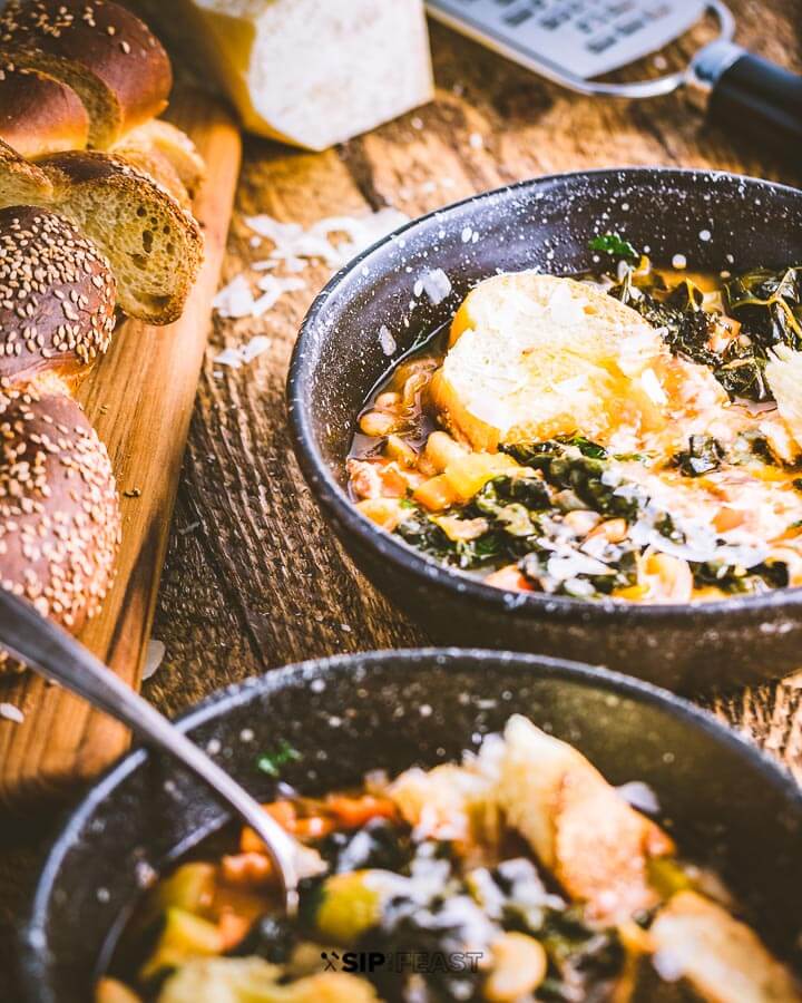 Two bowls of ribollita soup, loaf of Italian bread and cheese on table.