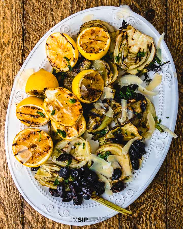 Grilled fennel and lemons with black olives and shaved cheese on white plate on wood table.