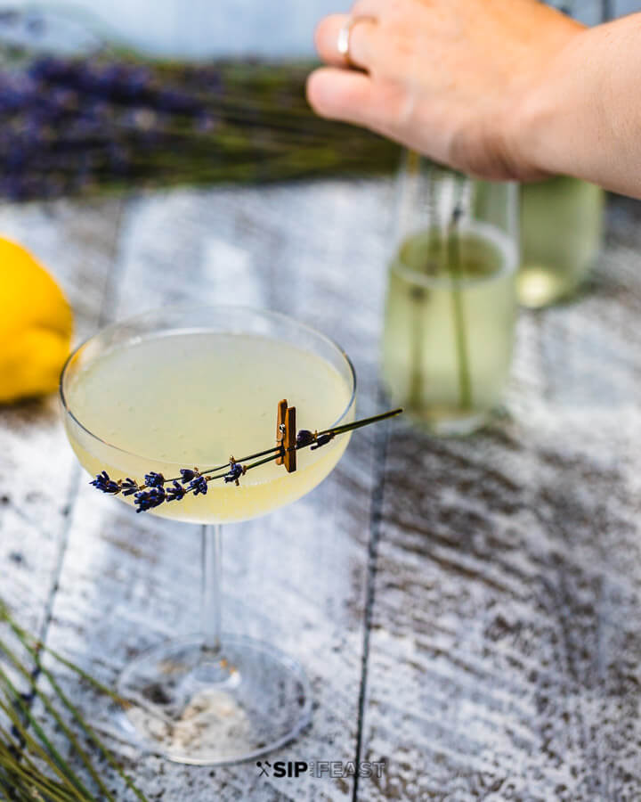 Glass of french 75 with lavender garnish and hands in background arranging more lavender in glasses.