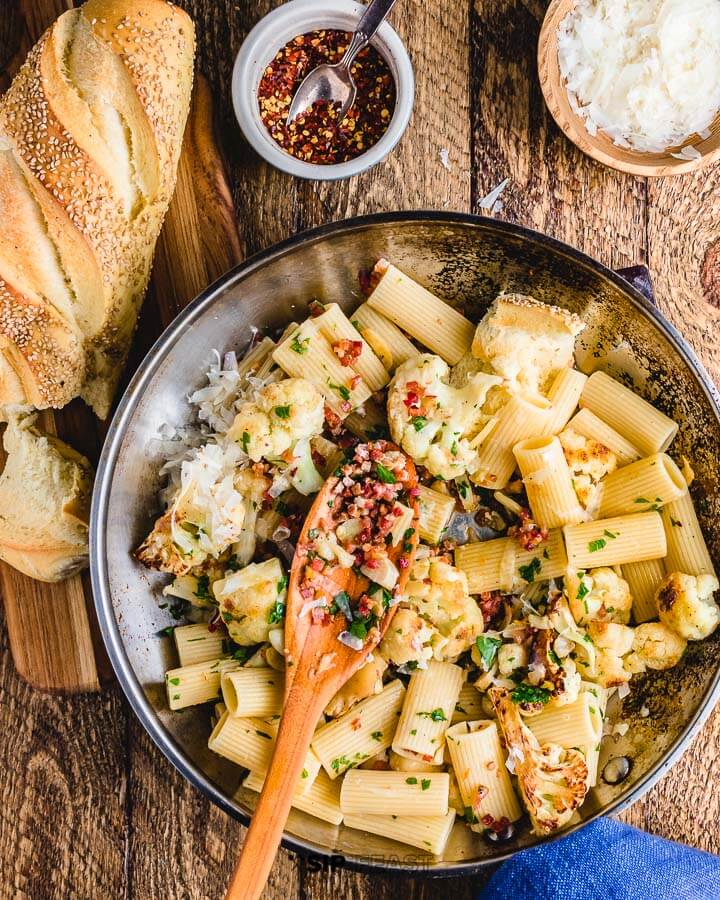 Rigatoni with cauliflower and bacon in large pan on wood table with cheese, bread and chili flakes on the side.