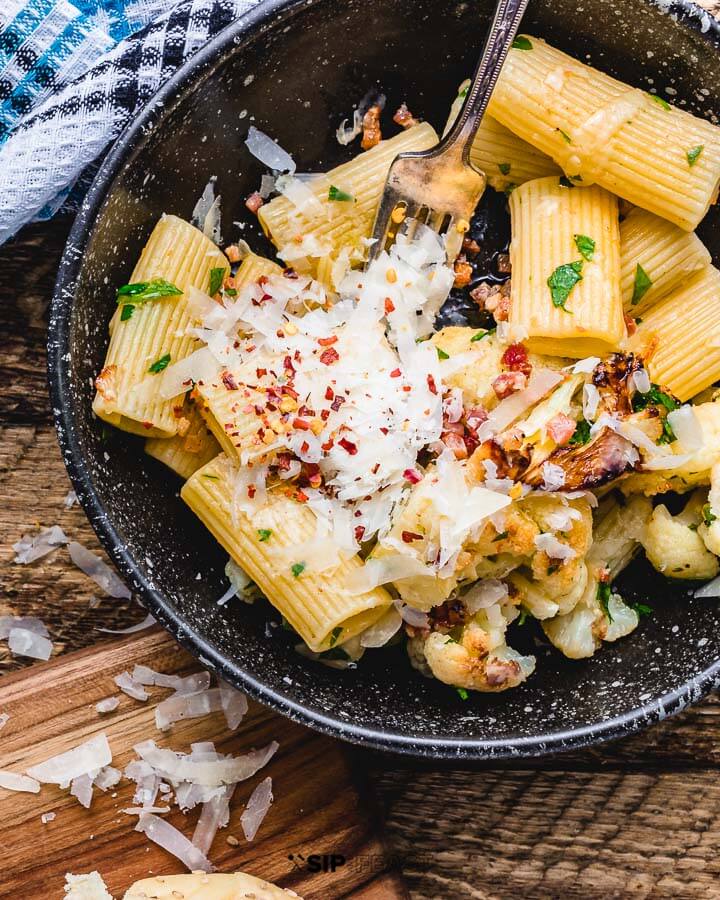 Rigatoni with cauliflower and bacon in black bowl on wood table.