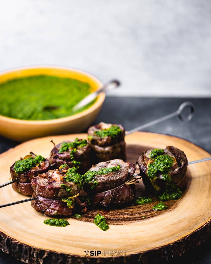 Skewered flank steak pinwheels on cutting board with bowl of green sauce on the side.