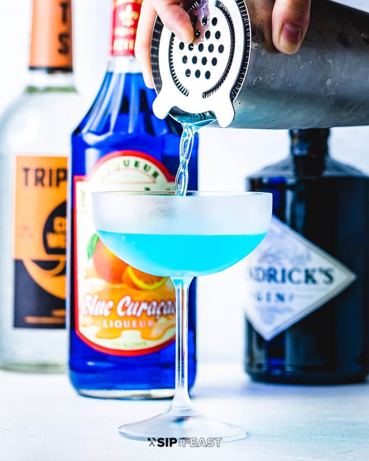 Cocktail shaker contents being poured into a coupe glass with bottles of gin, blue curacao and triple sec in the background.