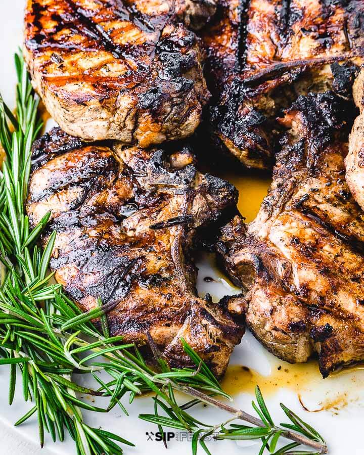 Four grilled pork chops in white plate with rosemary garnish.