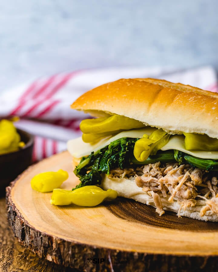 Sandwich with pulled Italian pork, broccoli rabe, provolone and Tuscan peppers on wood table.