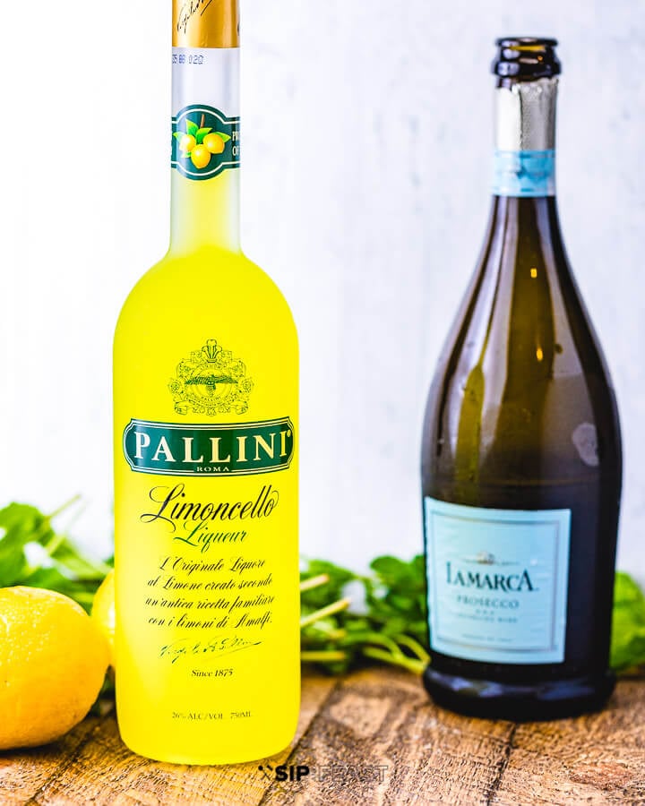 A bottle of limoncello and a bottle of prosecco with lemons and mint in the background.