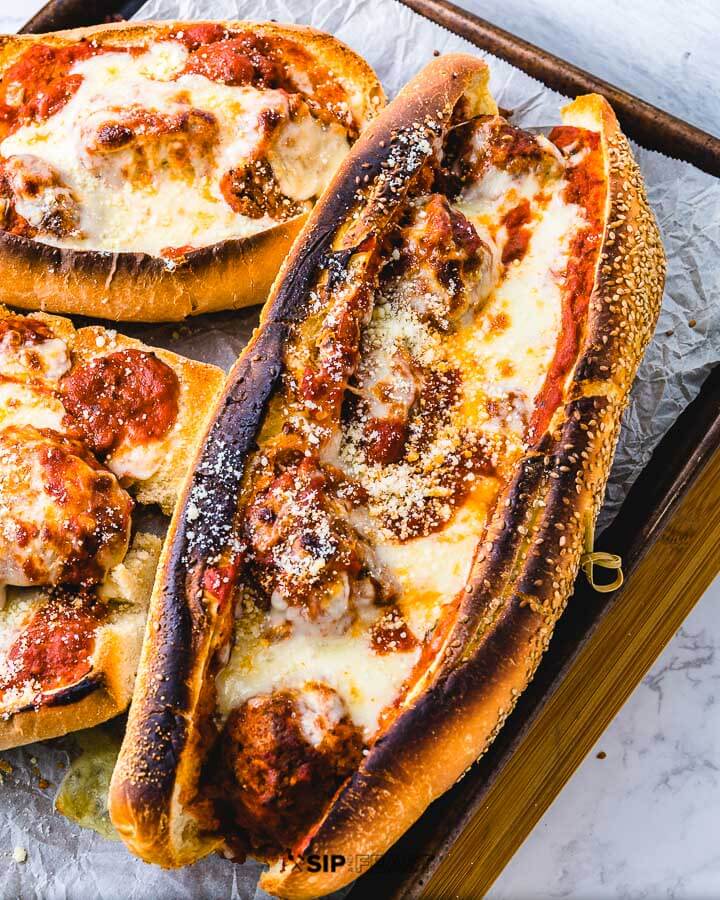 Three meatball parm heroes in a sheet pan on cutting board.