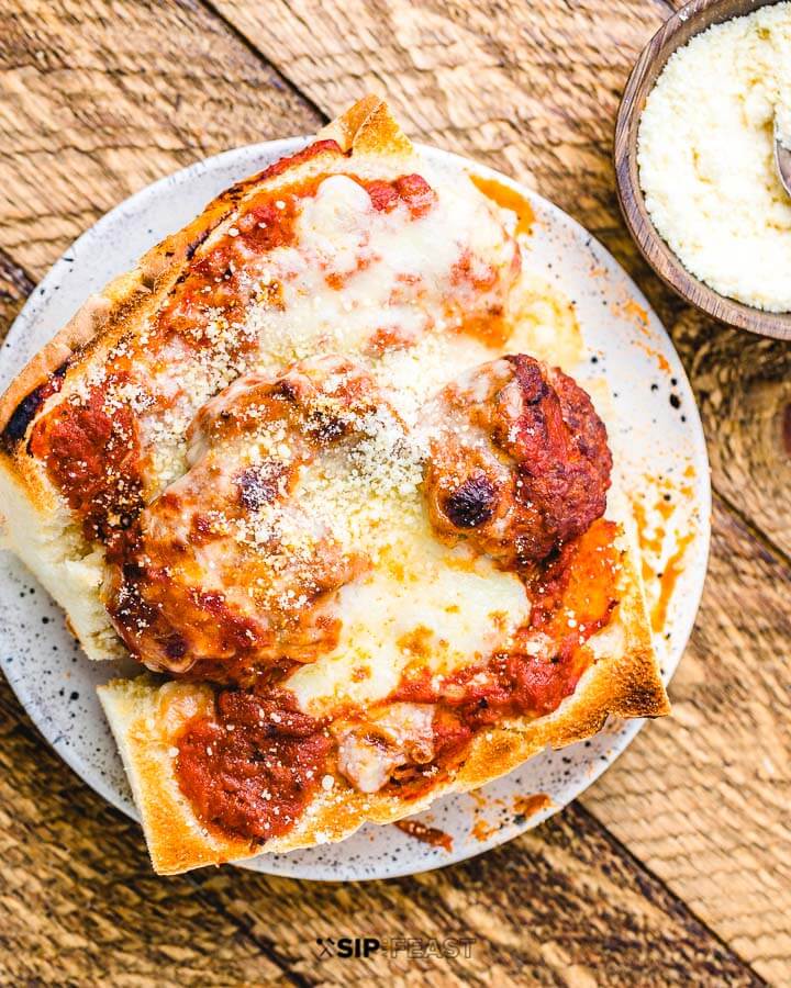 Small meatball sub on white plate with bowl of parmesan cheese on wood table.