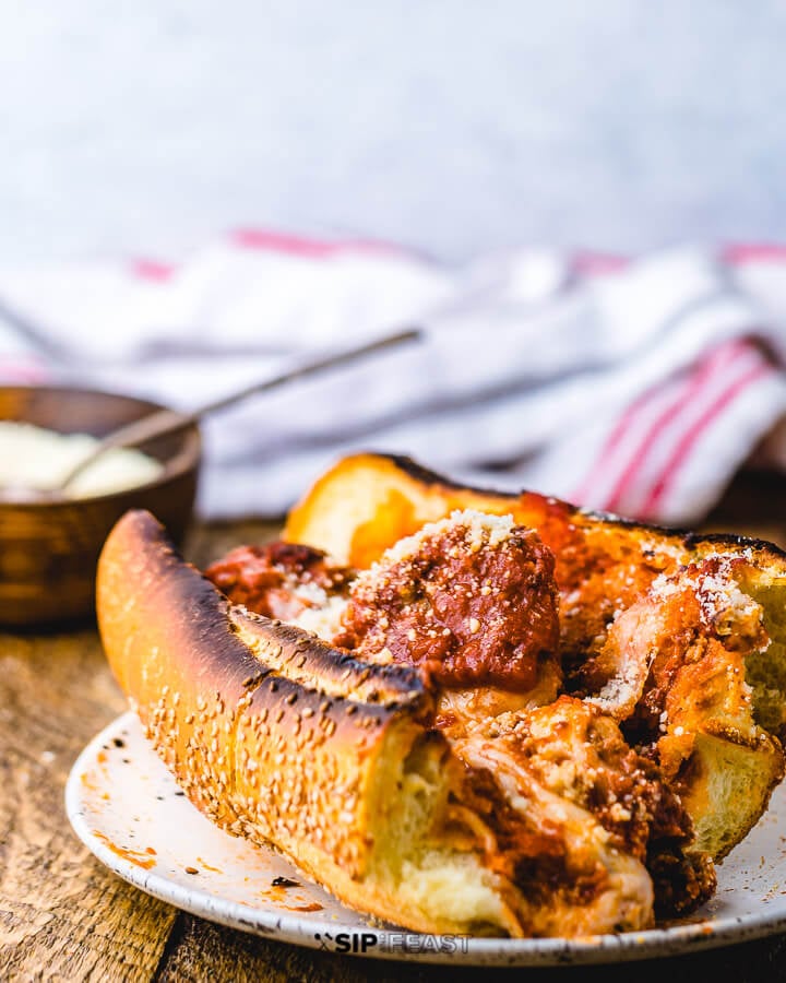 One half of a meatball sub in white plate on wood table with a bowl of parmesan.