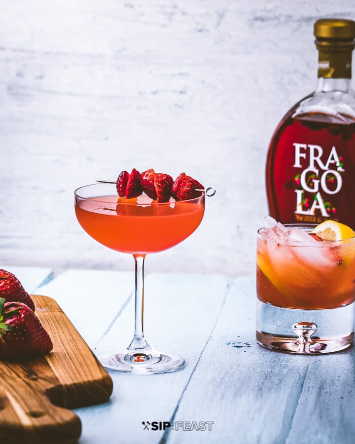 Cutting board with strawberries, two glasses of strawberry lemonade vodka cocktail, and bottle of Fragola liqueur.