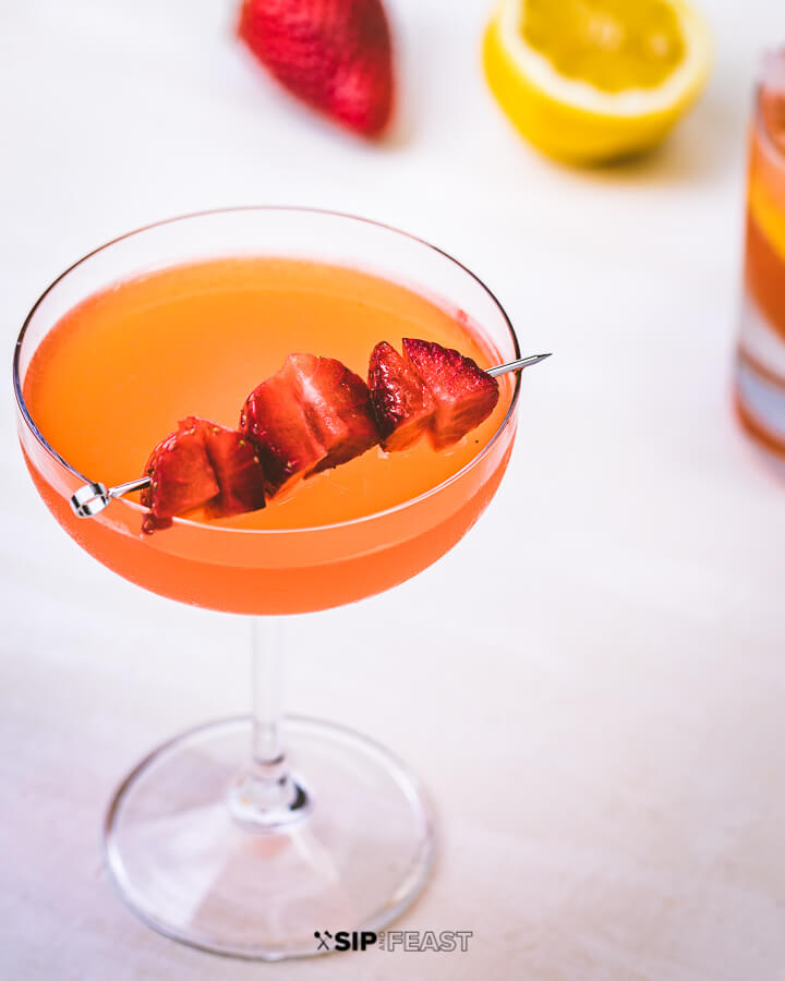 Glass of strawberry lemonade vodka with strawberry garnish, lemon and strawberry in background.