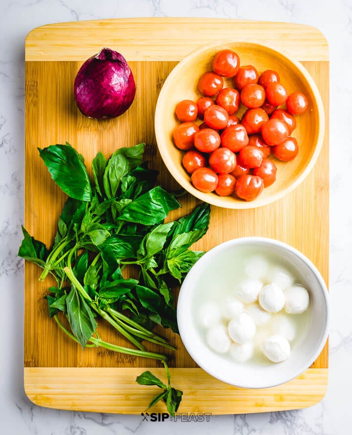 Ingredients on cutting board: red onion, grape tomatoes, basil and a bowl of fresh mozzarella.