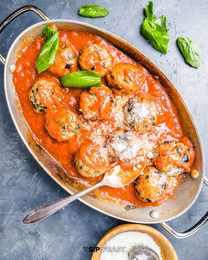 Eggplant meatballs in oval pan with sprinkled basil leaves and bowl of Pecorino Romano cheese.