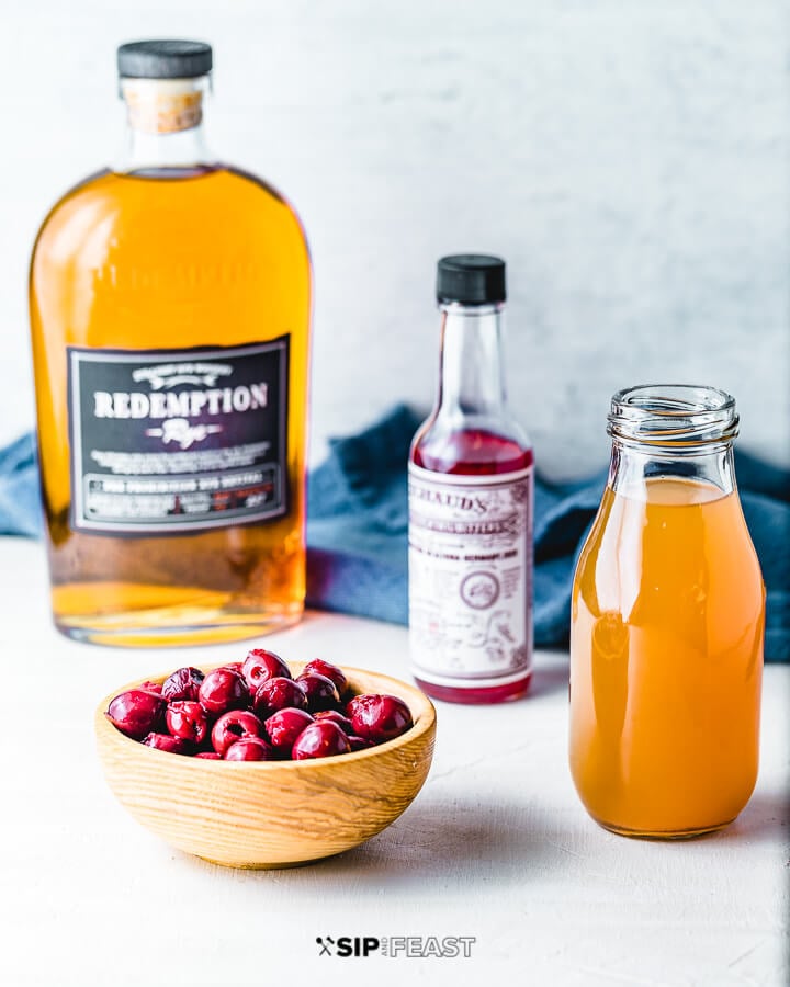 A bottle of rye, cocktail bitters, a bowl of cherries and apple cider.