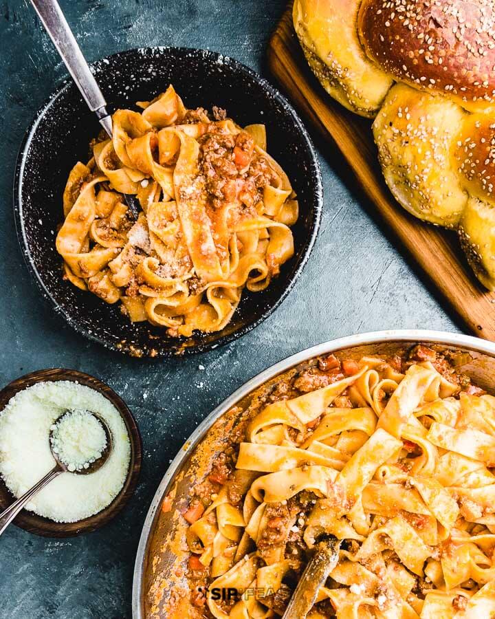 Large pan and small bowl of pappardelle bolognese on blue background.