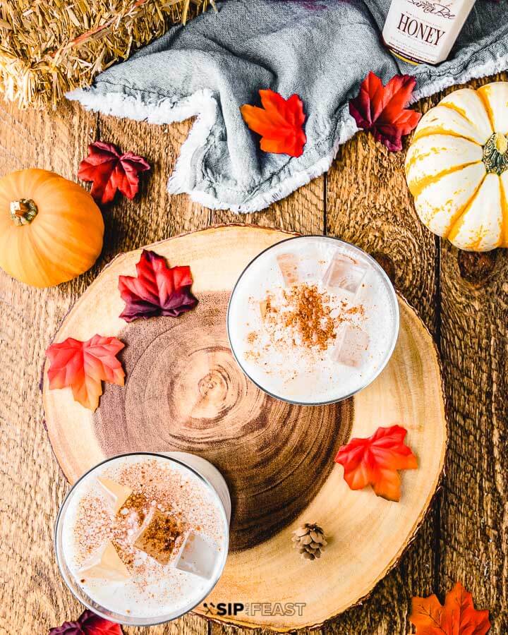 Two glasses with pumpkin spice bourbon milk punch, decorative leaves and pumpkins on a table top.