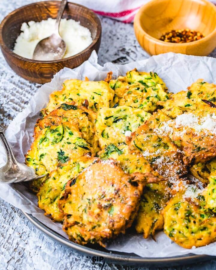 Zucchini fritters in large plate with bowl of Pecorino Romano cheese and crushed red pepper flakes in background.