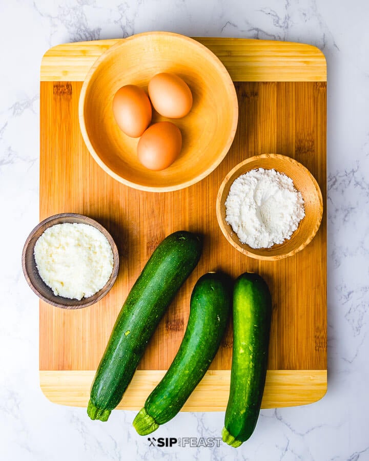 Ingredients shown: eggs in bowl, flour in bowl, cheese in bowl and 3 zucchini.