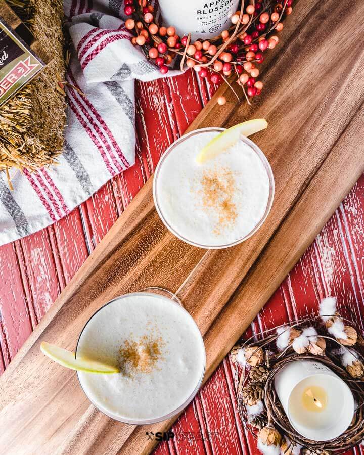 Two coupe glasses filled with apple pie martini with Fall scape in background that includes candles, hay bales and berries.