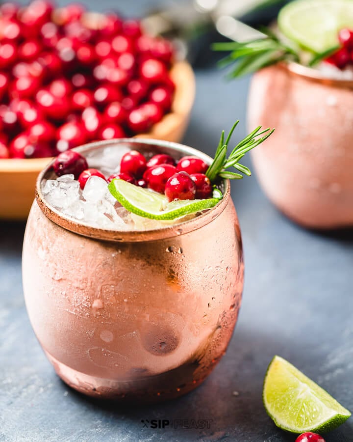 Two copper mugs with the Cranberry Moscow Mule cocktail, and a bowl of cranberries.