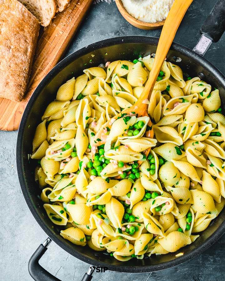 Large pan of shells with pancetta and peas on blue background.