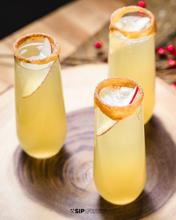 3 caramel apple mimosas in stemless champagne flutes garnished with a rim of caramel, sugar and cinnamon, and one slice of apple.