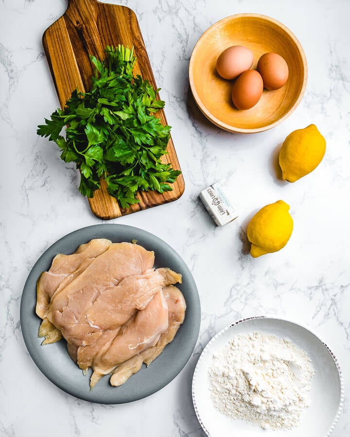 Ingredients shown: parsley, eggs, butter, lemons, chicken cutlets, and bowl of flour.