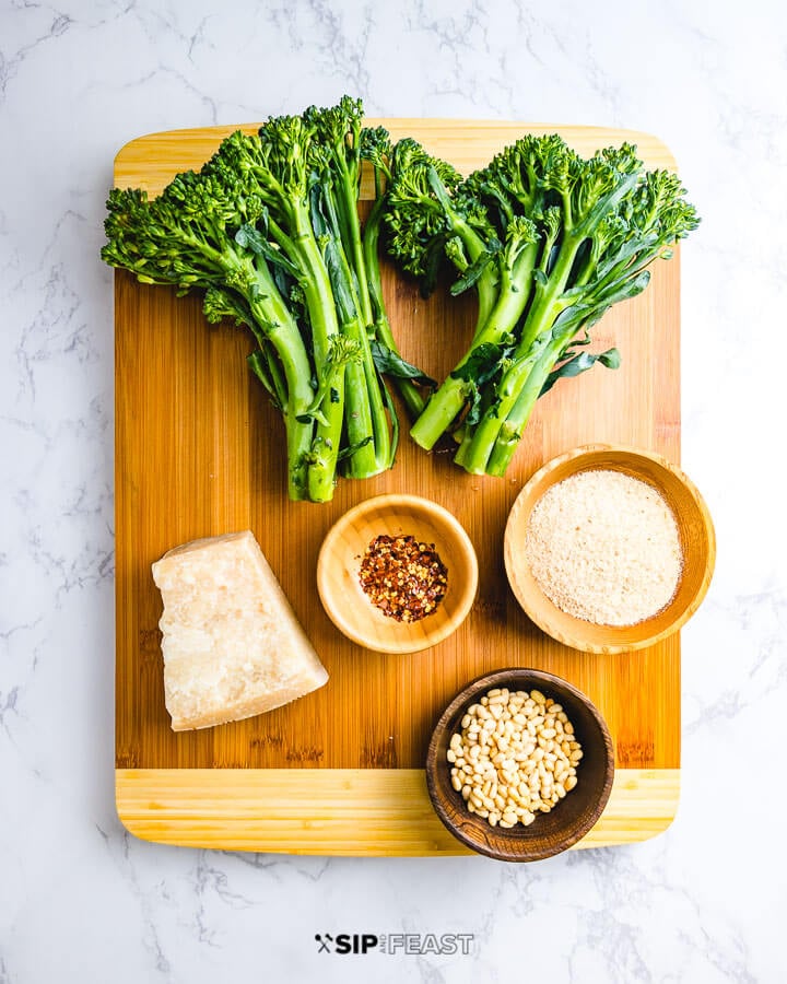 Ingredients shown on cutting board: baby broccoli, parmesan cheese, pine nuts, breadcrumbs, and chili flakes.