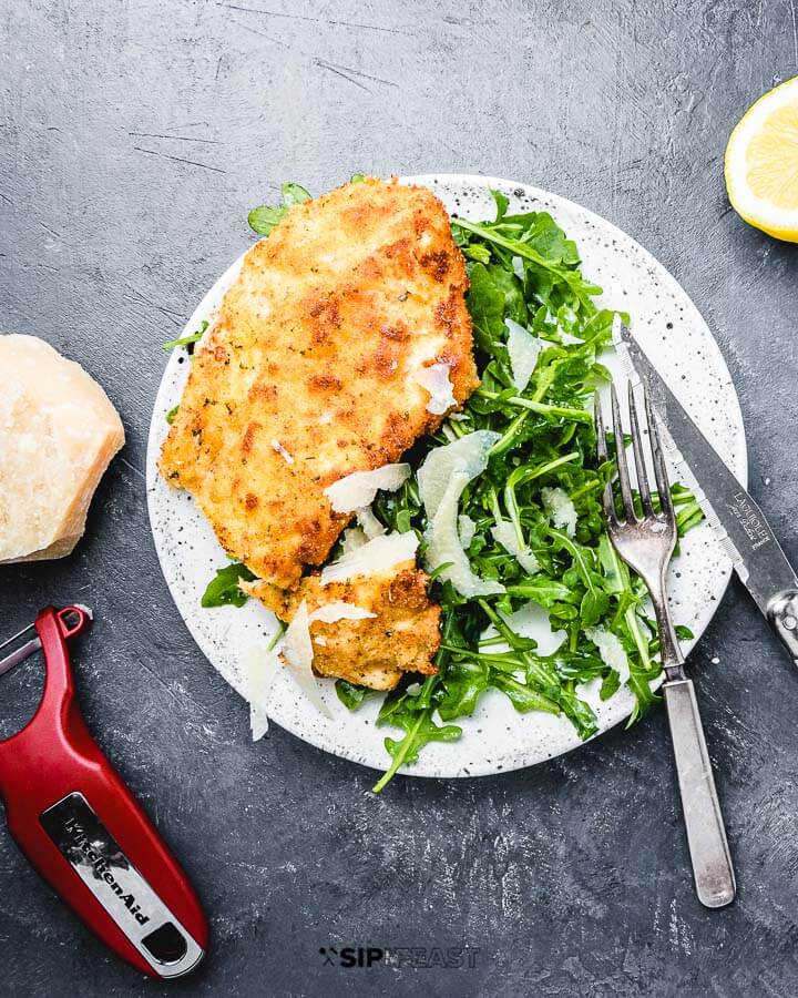 Chicken Milanese in white plate with lemon and parmesan cheese on the side.