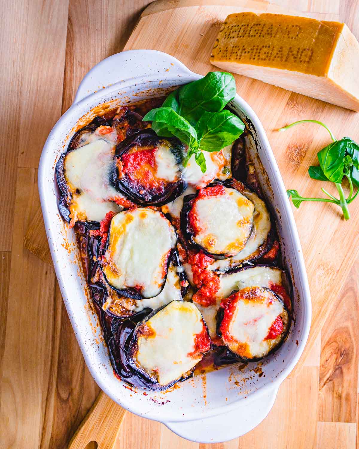 Overhead shot of large dish with finished eggplant parmesan and block of Parmigiano Reggiano cheese.