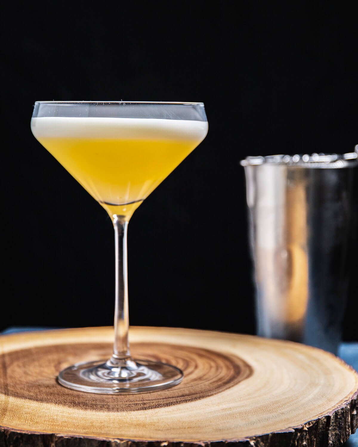 Oliveto cocktail in coupe glass on round wooden board with metal shaker in background.