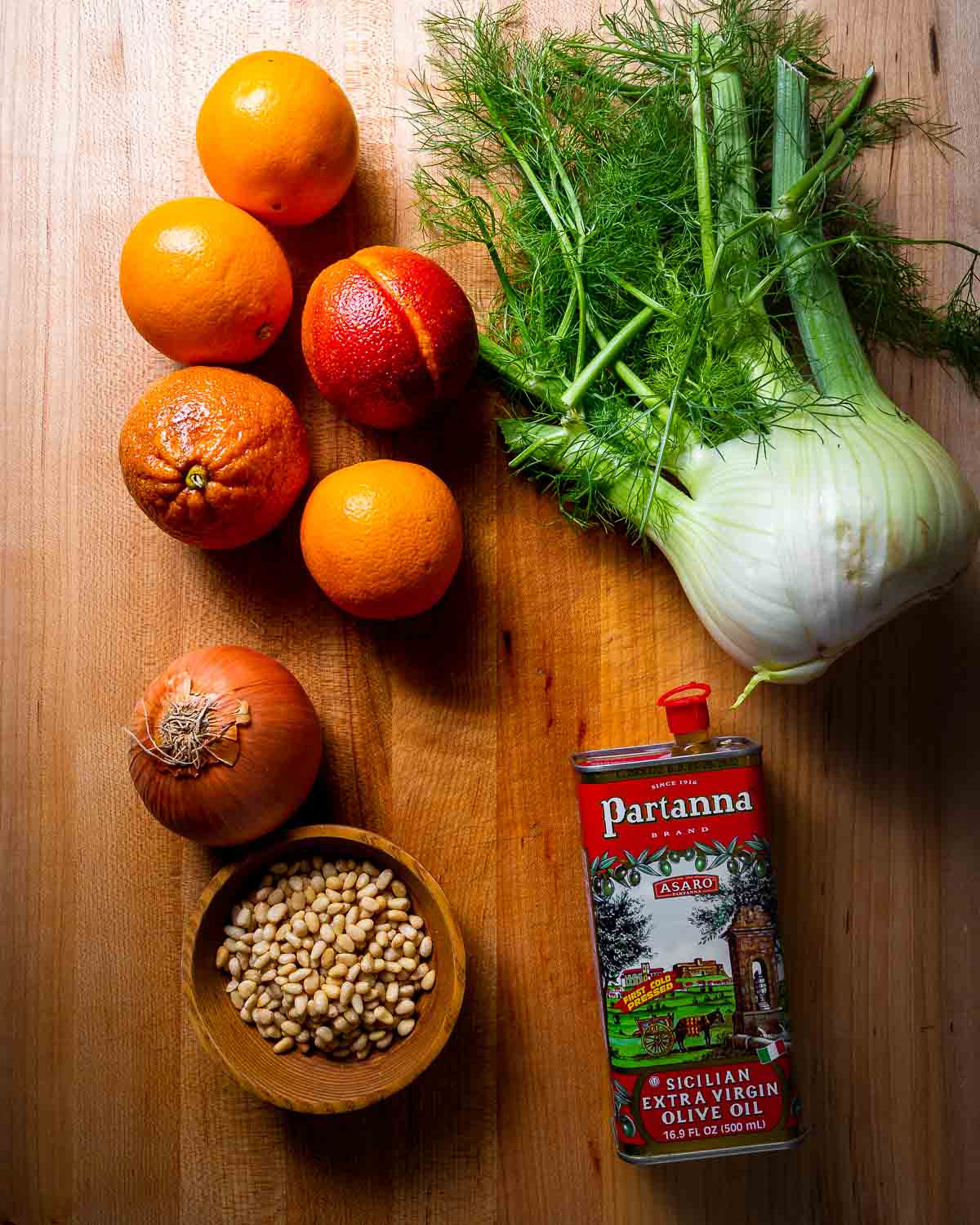 Ingredients shown: 5 oranges, 1 onion, 1 fennel, pine nuts and extra virgin olive oil on a wood cutting board.