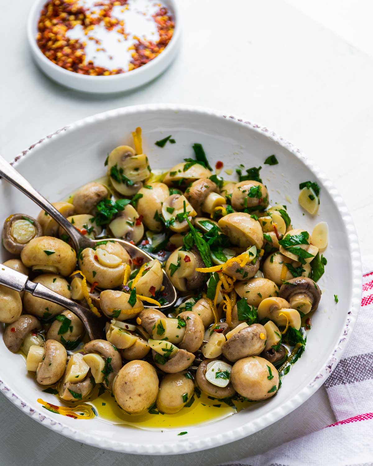 Marinated mushrooms in white bowl with spoon and fork on white table.