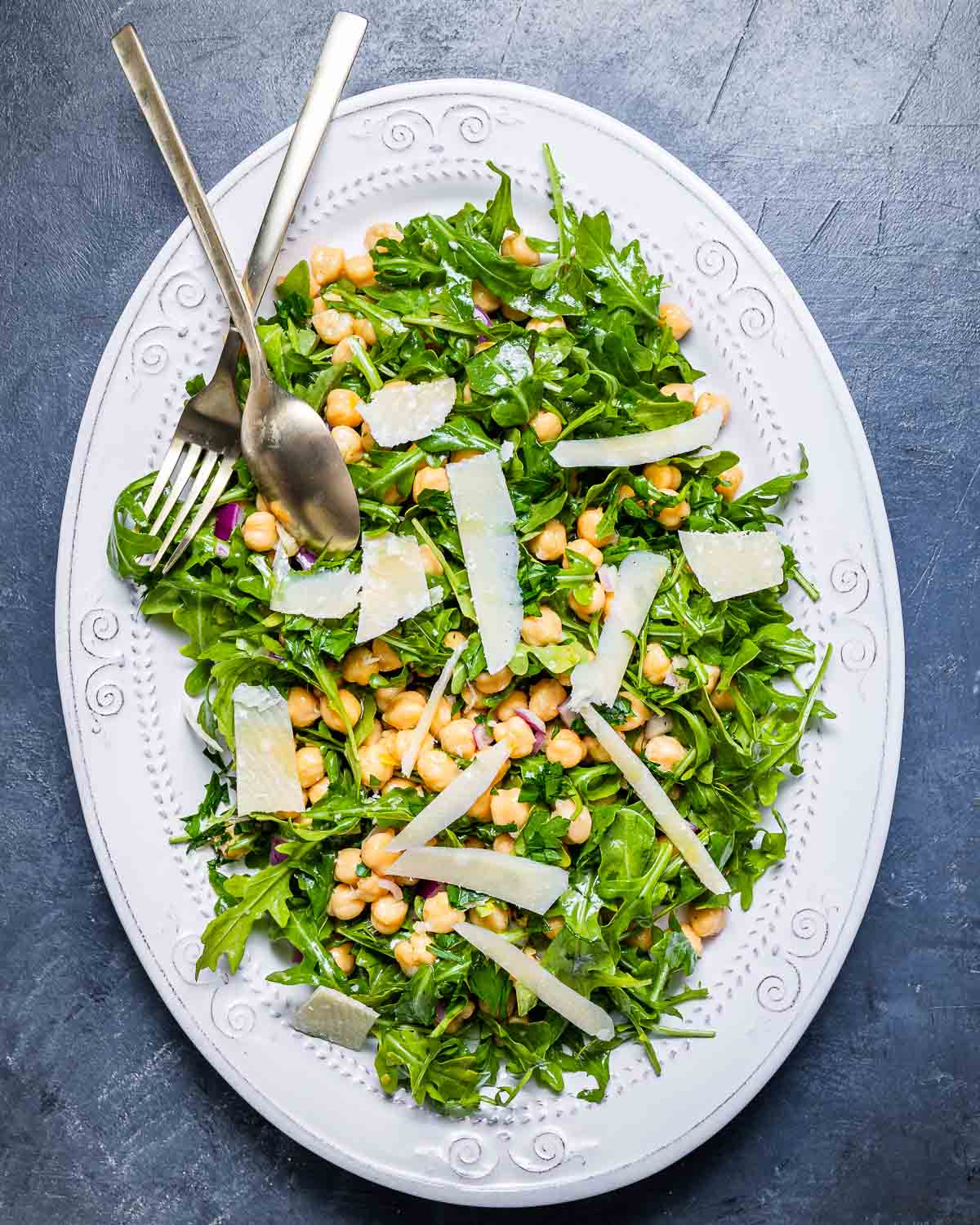 Overhead shot of white platter with arugula, chickpeas, and shaved cheese on blue board.