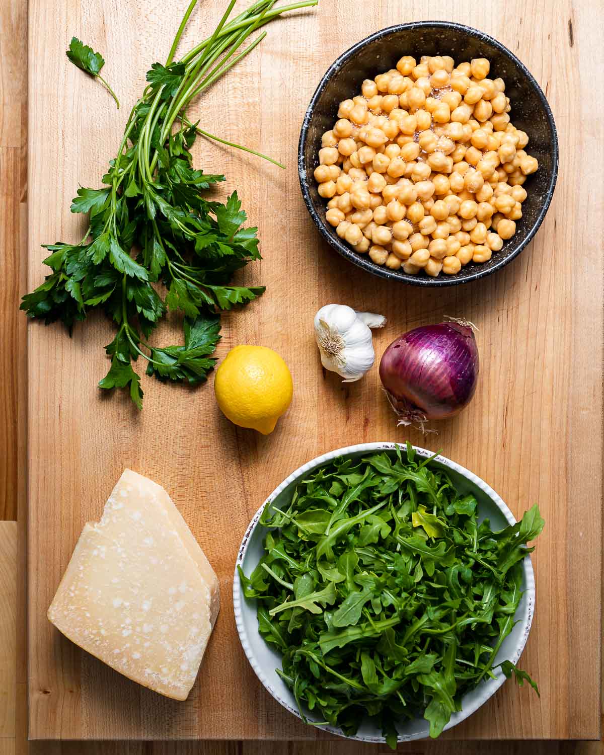 Ingredients shown: parsley, chickpeas, lemon, garlic, red onion, block of parmesan, and bowl of arugula.