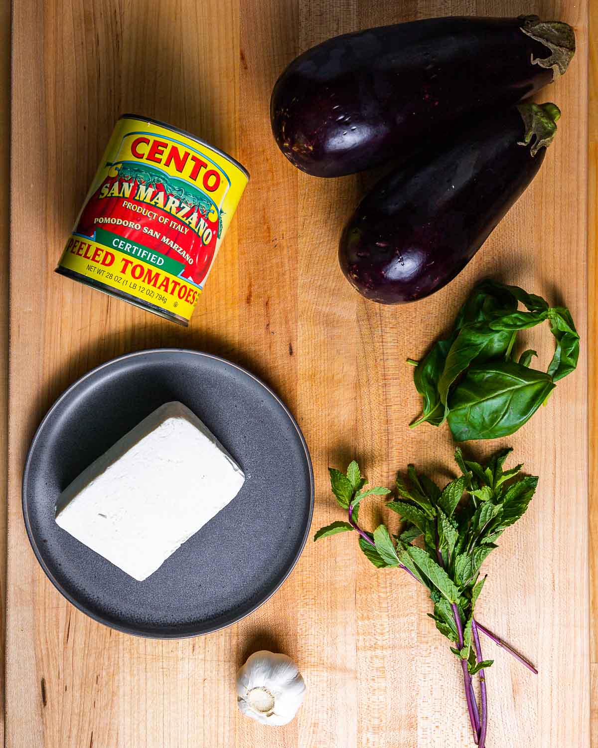 Ingredients shown: Can of plum tomatoes, 2 eggplants, ricotta salata, garlic, mint and basil.