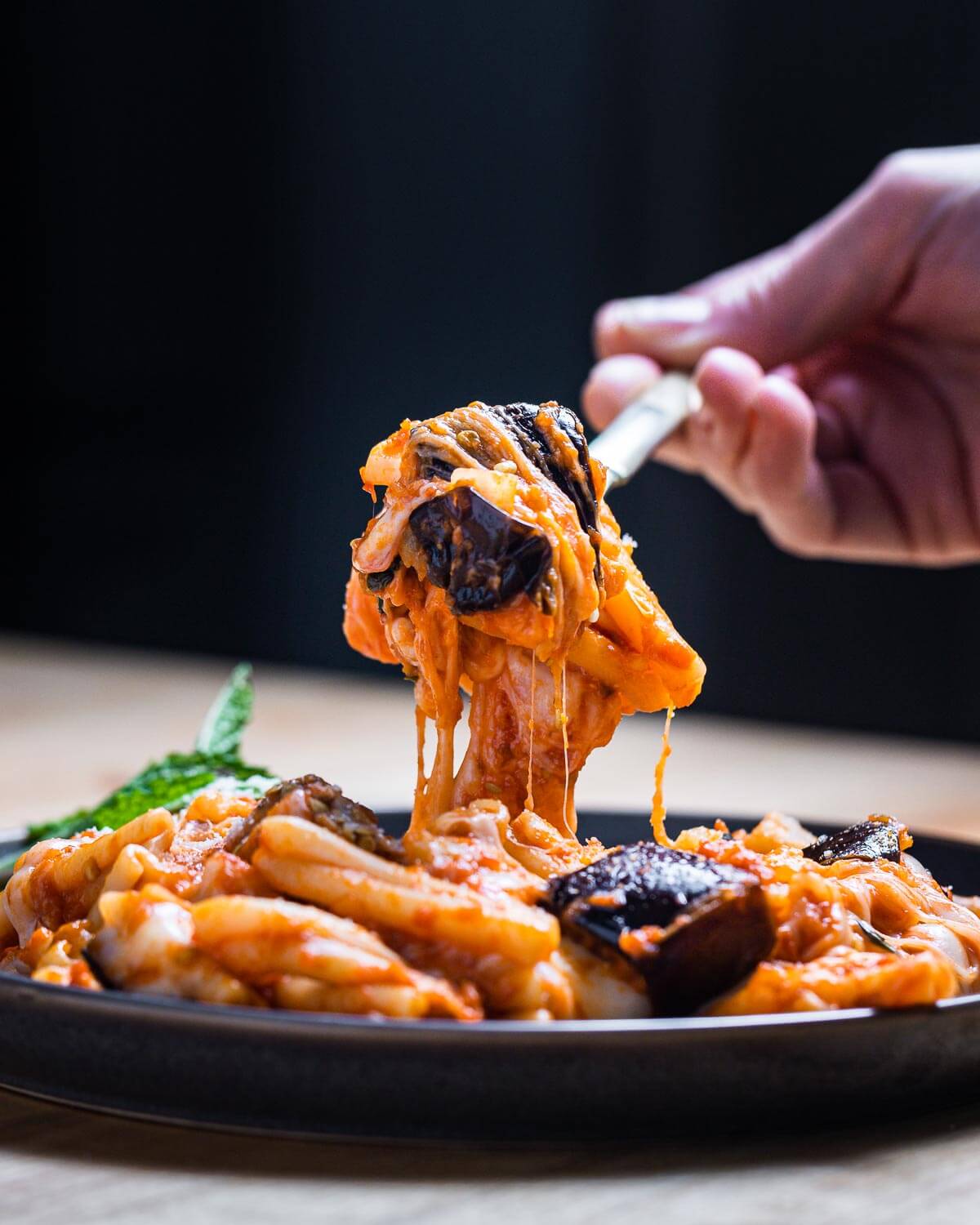 Pasta, eggplant, and mozzarella held in air with fork over plate of Pasta alla Siciliana.