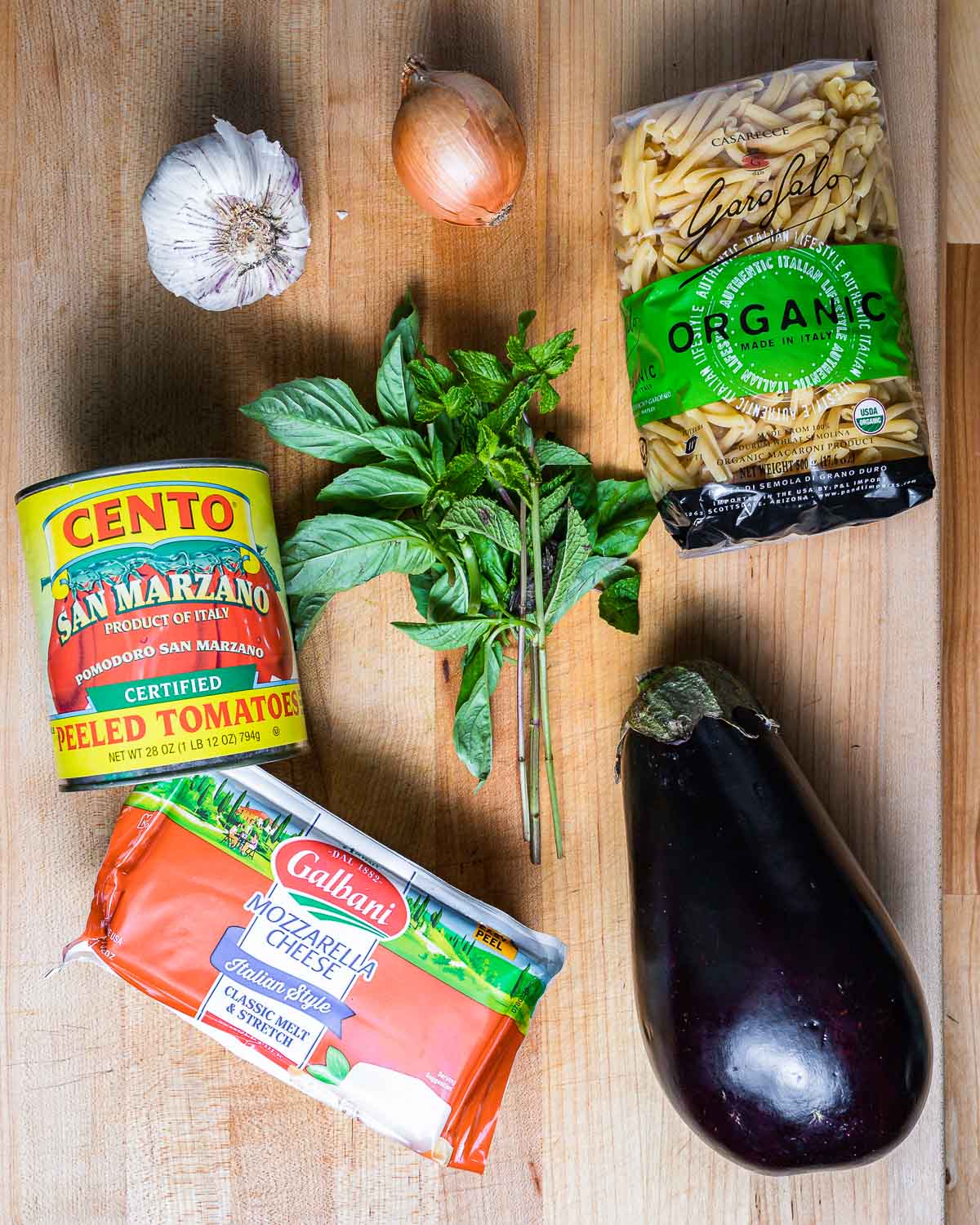 Ingredients shown: garlic, onion, casarecce pasta, plum tomatoes, basil, mint, mozzarella, and large eggplant.