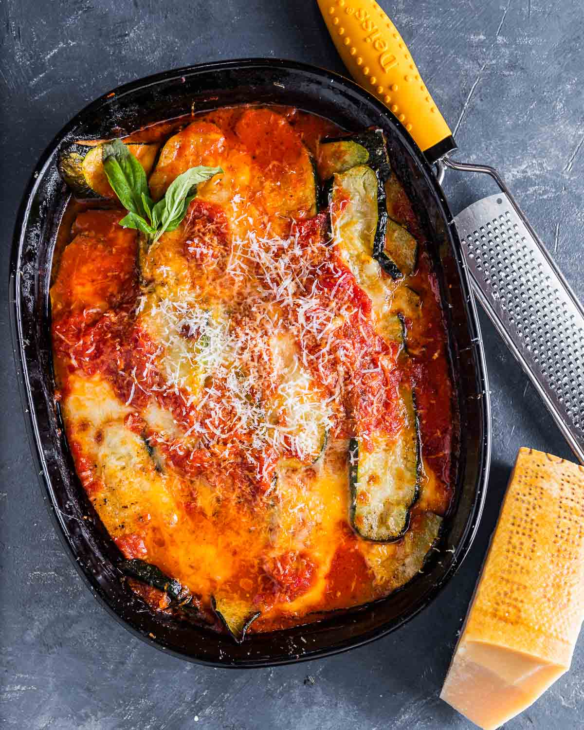 Overhead shot of baked zucchini parm in baking dish with block of Parmigiano Reggiano cheese on the side.