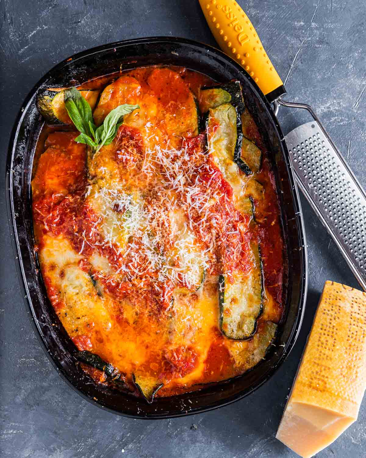 Overhead shot of baked zucchini parm in baking dish with block of Parmigiano Reggiano cheese on the side.