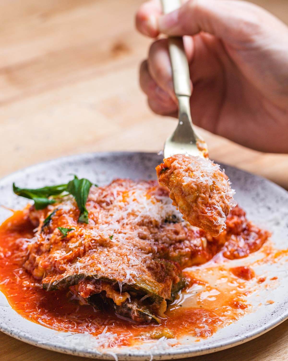 Fork holding piece of zucchini parm over white plate.