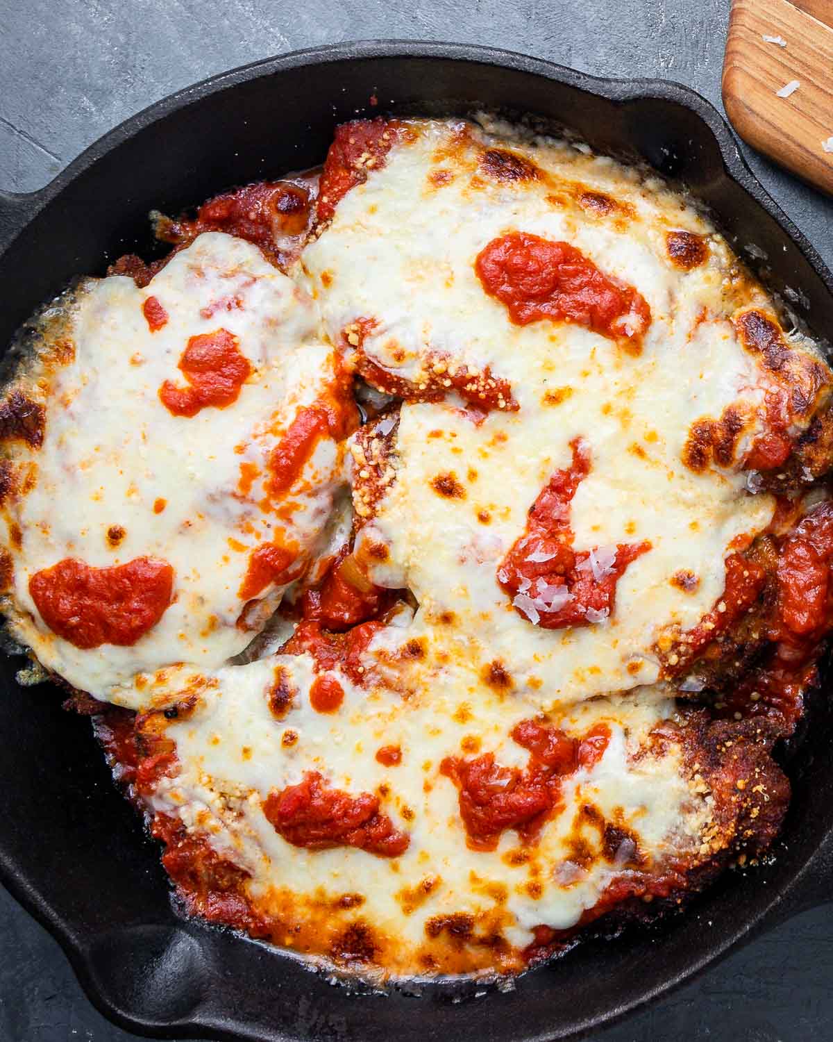 Overhead shot of cooked chicken parm in cast iron pan on blue board.