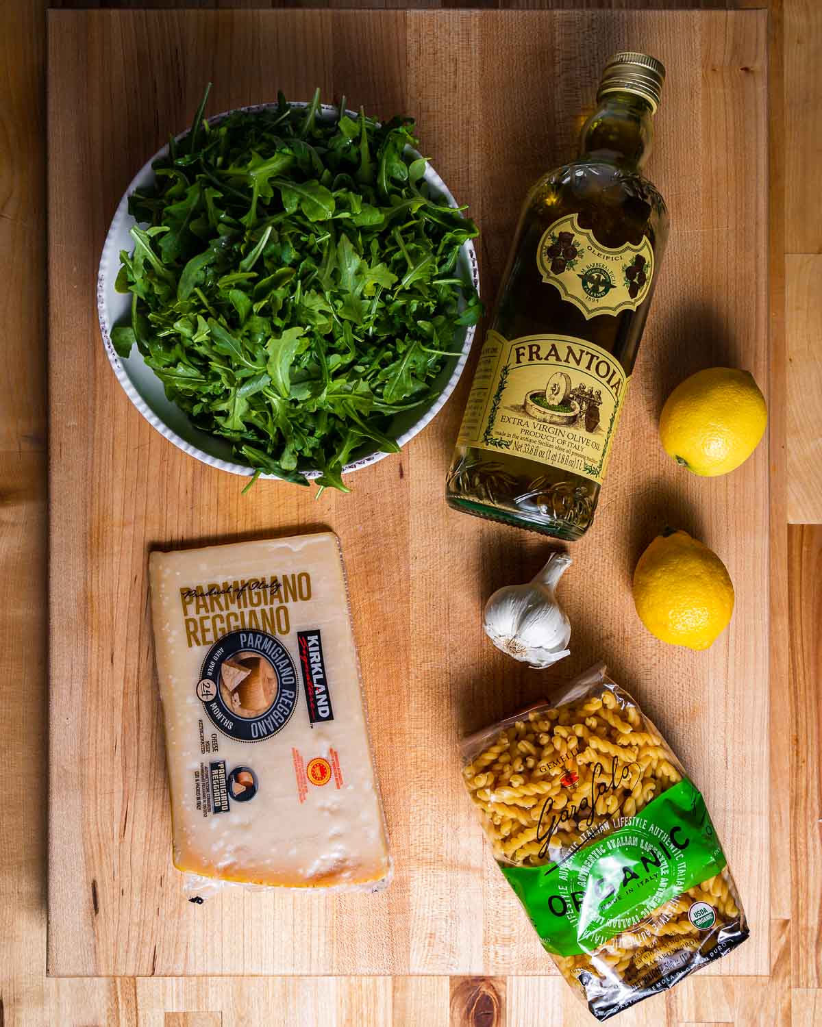 Ingredients shown: bowl of arugula, olive oil, parmesan cheese, lemon, garlic, and pasta.