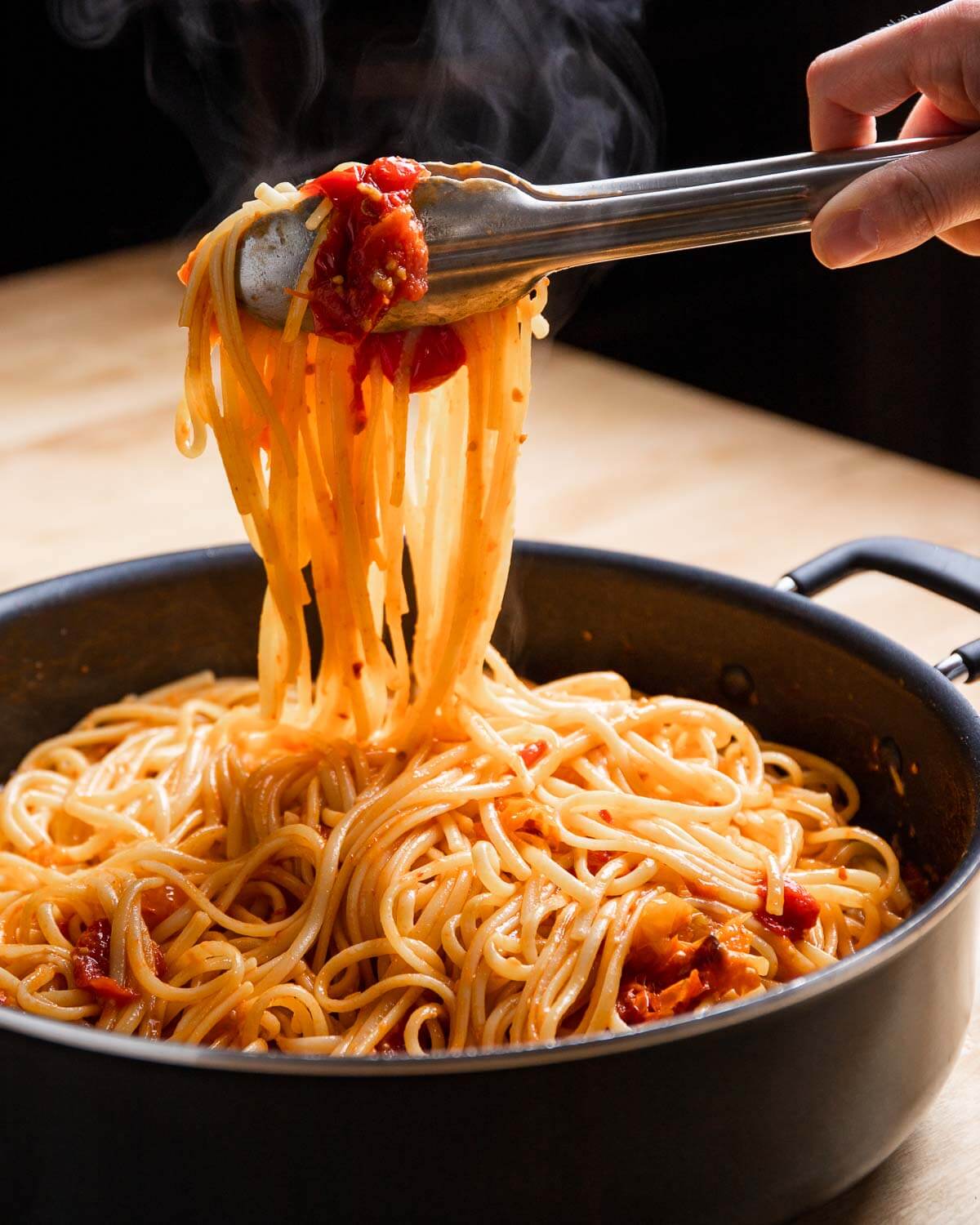 Tongs holding linguine with cherry tomato sauce over large pan.