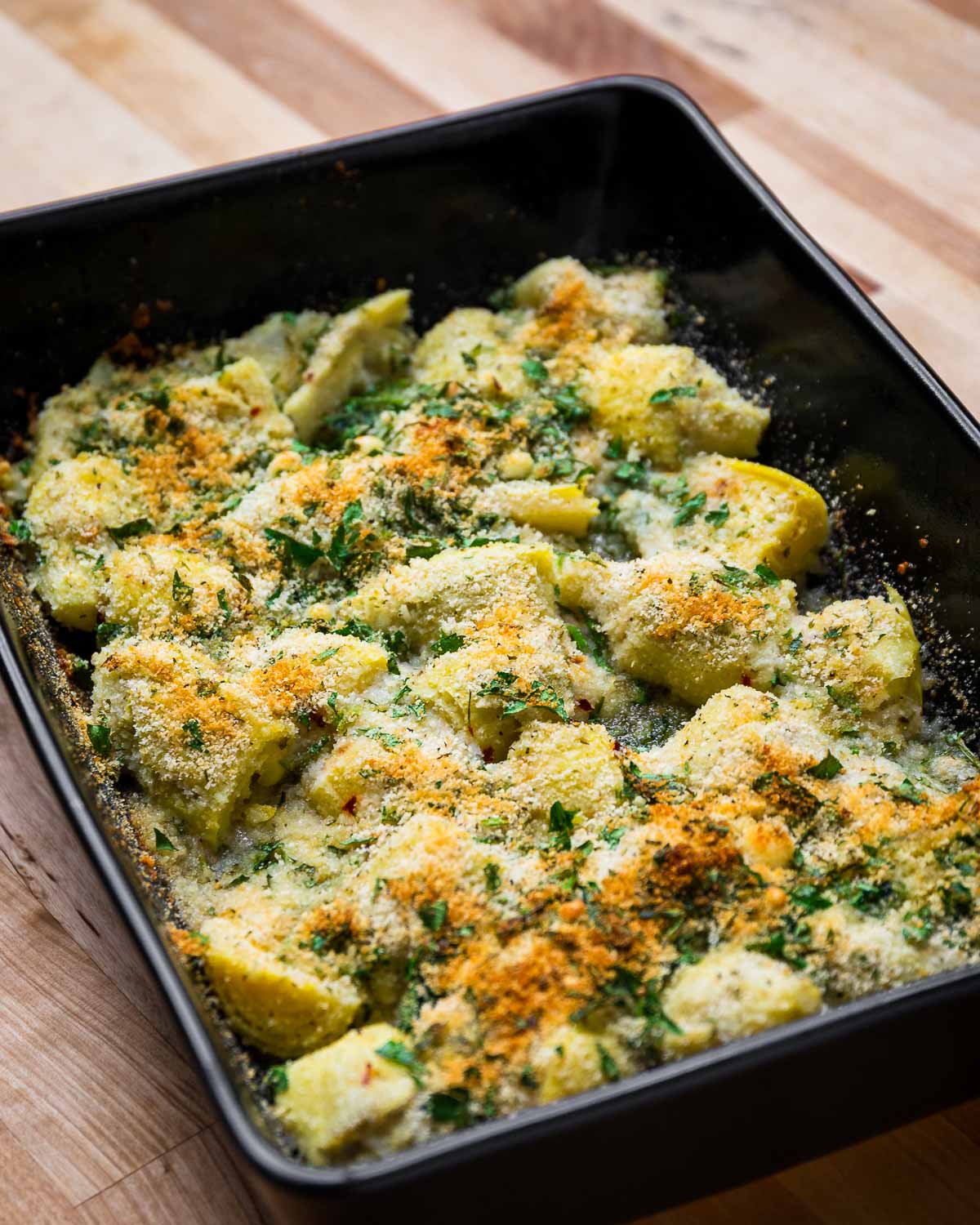 Baked artichoke hearts in black baking dish on wood table.