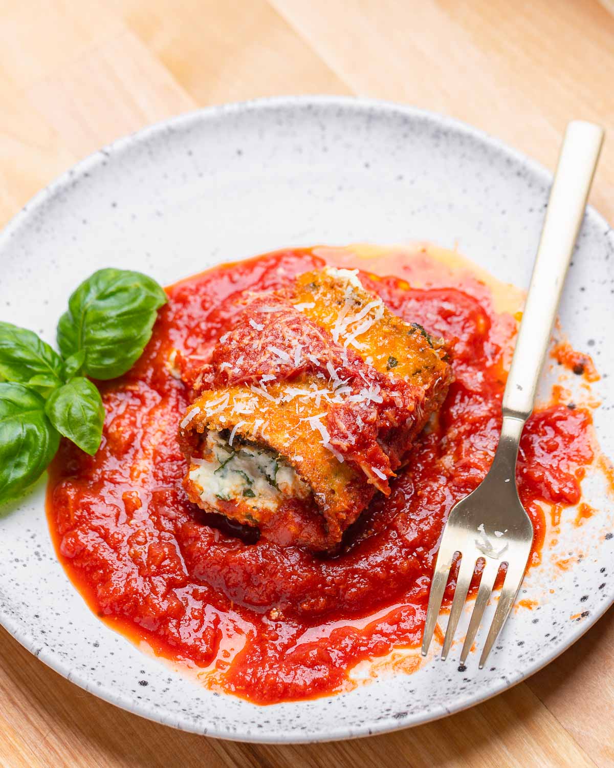 White plate with one eggplant rollatini, basil leaves, and fork.